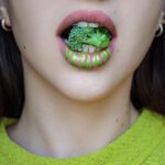 A close-up of a person's open mouth filled with broccoli, with the word "vegan" written on their lips to market to non-vegans.