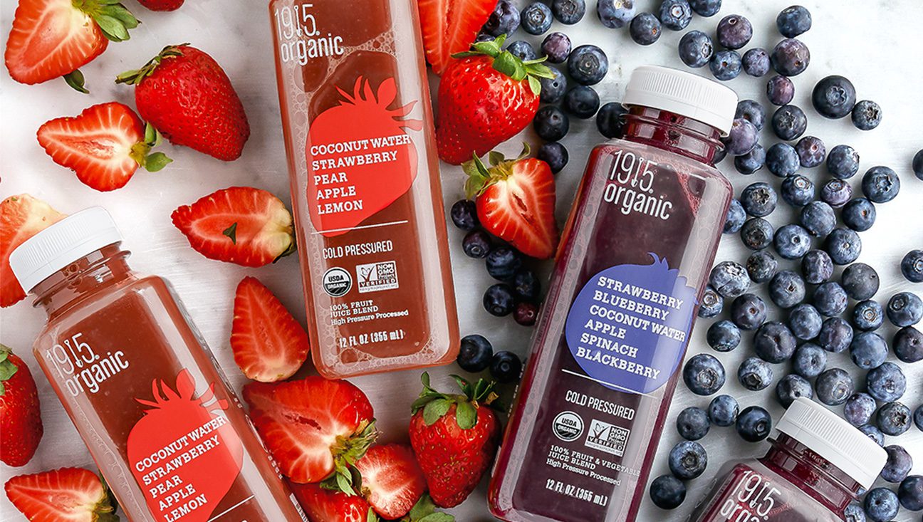 Three bottles of 1915 Organic cold-pressed juice surrounded by fresh strawberries and blueberries.