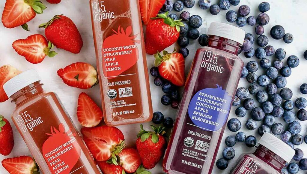 Several bottles of 1915 Organic cold-pressed juices are surrounded by fresh strawberries and blueberries. The flavors shown include Coconut Water Strawberry Pear Apple Lemon and Strawberry Blueberry Coconut Water Apple Spinach Blackberry. Perfect for direct-to-consumers, these vibrant juices are a hit on social media.