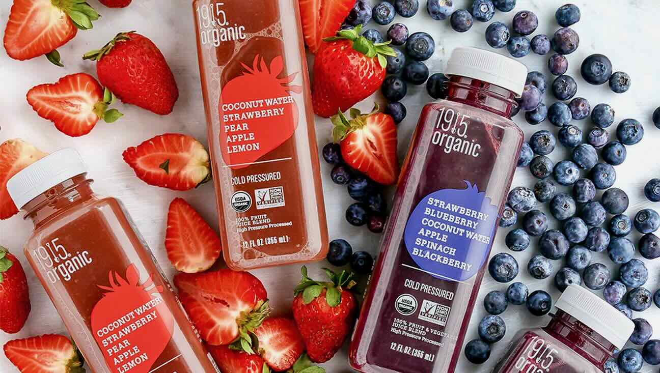 Several bottles of 1915 Organic cold-pressed juices are surrounded by fresh strawberries and blueberries. The flavors shown include Coconut Water Strawberry Pear Apple Lemon and Strawberry Blueberry Coconut Water Apple Spinach Blackberry. Perfect for direct-to-consumers, these vibrant juices are a hit on social media.
