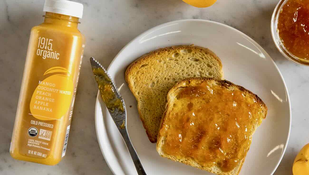 A bottle of 1915 Organic juice next to a plate with a knife, a piece of toast topped with orange jam, another toast, and a small bowl of jam sets the perfect scene for showcasing our B2C marketing approach on social media strategies.