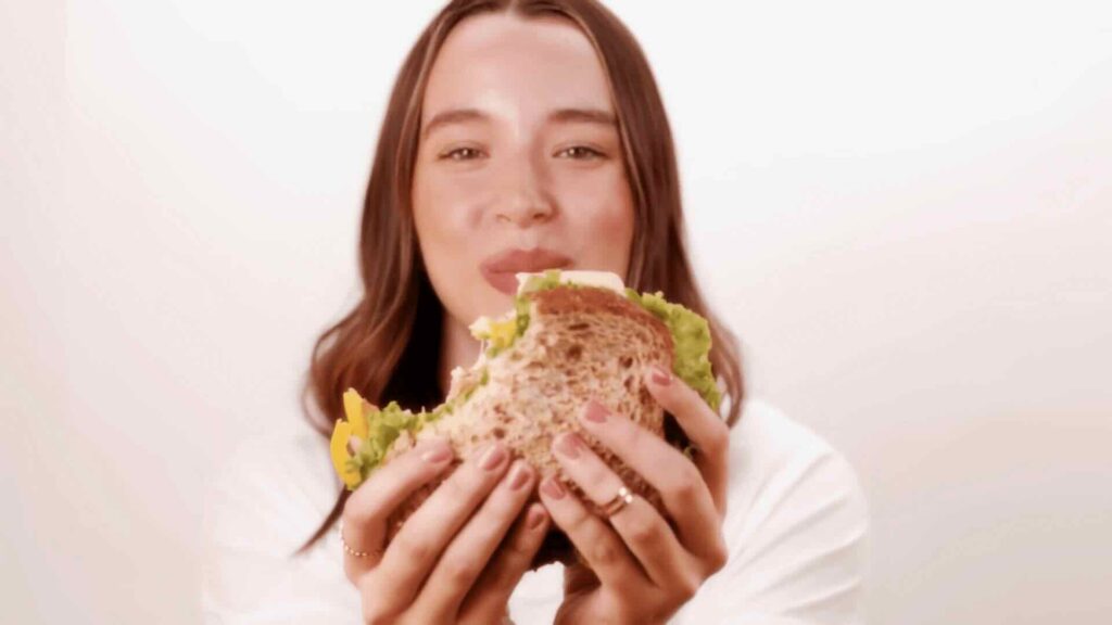 A person with long hair smiles and holds an Angelic Bakehouse sandwich with multigrain bread, lettuce, and other fillings up to their mouth, ready to take a bite. This moment is part of the #BreadBod campaign's hero videos for our social media strategy.