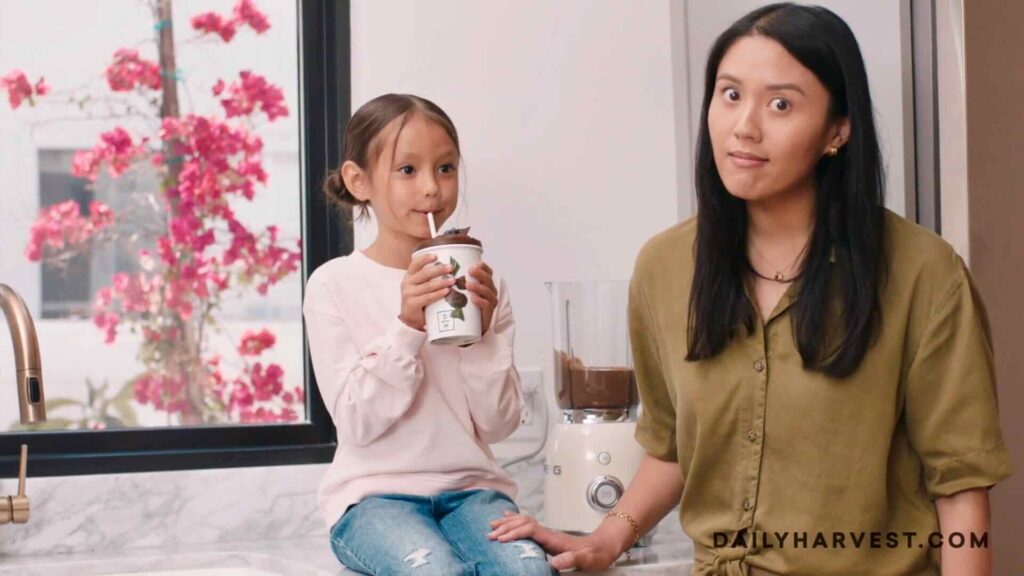 A woman stands in a kitchen next to a young girl who is sitting on the counter, drinking from a cup with a straw. The letters "DAILY HARVEST," part of their content marketing strategy, are visible in the bottom right corner.