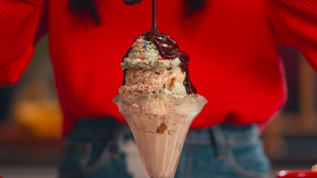 A dad, part of the Babyganics Open for Adventure campaign, is pouring chocolate syrup over a multi-flavored ice cream sundae in a glass dish, wearing a red sweater and jeans. This moment captures the essence of family joy and strategic partnerships through digital platforms.