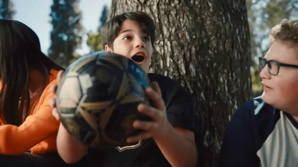 A child holding a soccer ball with an excited expression sits between two other children near a tree, embodying the joyful spirit often highlighted in content marketing campaigns.