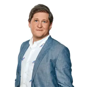 A person with short brown hair wearing a light blue blazer and a white shirt stands against a plain white background.