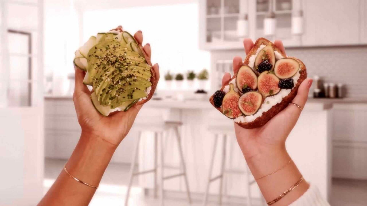 Two hands hold open-faced sandwiches: one topped with avocado slices and seasonings, the other with figs, blackberries, and a creamy spread. This delectable scene features Angelic Bakehouse #BreadBod campaign in a bright, white kitchen background.
