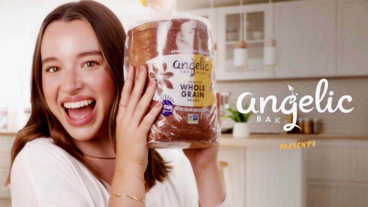A smiling woman in a kitchen holds up a loaf of Angelic Bakehouse Whole Grain bread while looking at the camera. As part of the Angelic Bakehouse #BreadBod campaign, the logo is prominently displayed on the right side of the image.
