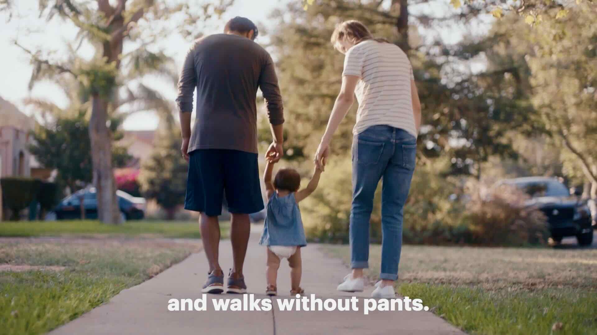 A man and a woman walk on a sidewalk holding hands with a small child in the middle. Text at the bottom reads, "and walks without pants," highlighting Babyganics' Open for Adventure campaign that celebrates adventurous parenting and family joy.