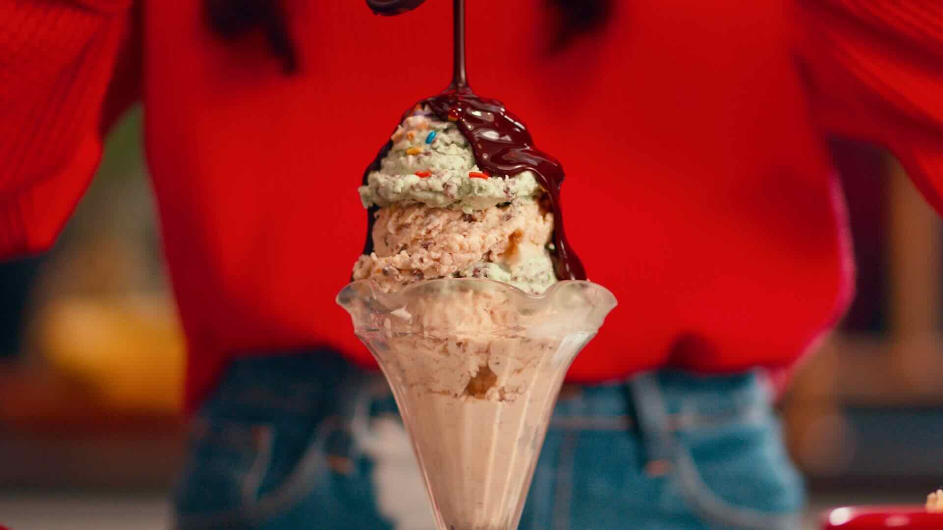 A person in a red sweater pours chocolate syrup over a tall ice cream sundae with multiple scoops in a glass dish, adding to the joy of family celebrations. It's moments like these that Stater Bros. Markets celebrates, showcasing their commitment to quality products and community in Southern California.