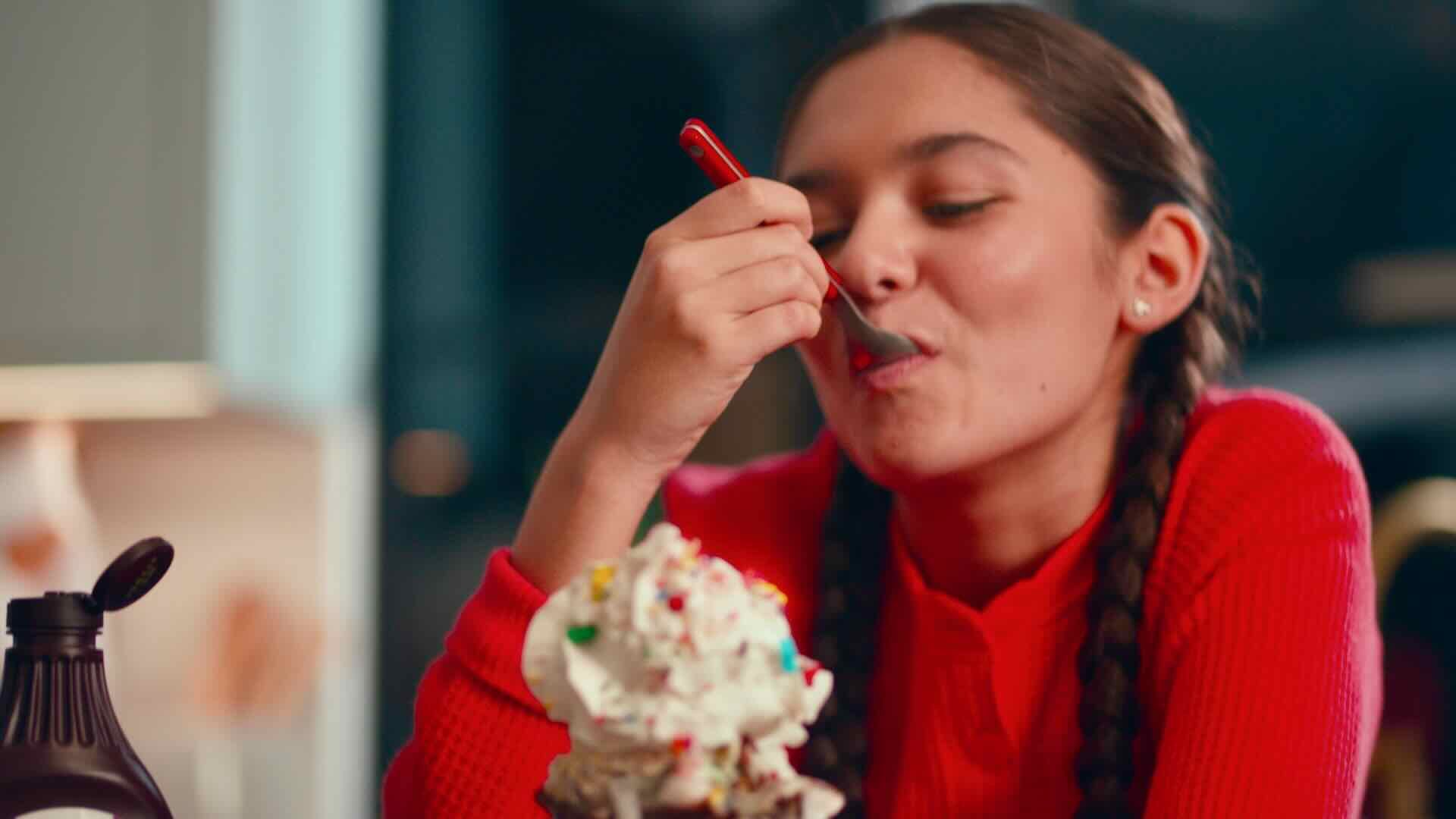 A person with braided hair, wearing a red sweater, enjoys a spoonful of dessert topped with whipped cream and sprinkles, celebrating a family milestone as seen in the Stater Bros. Markets commercial.