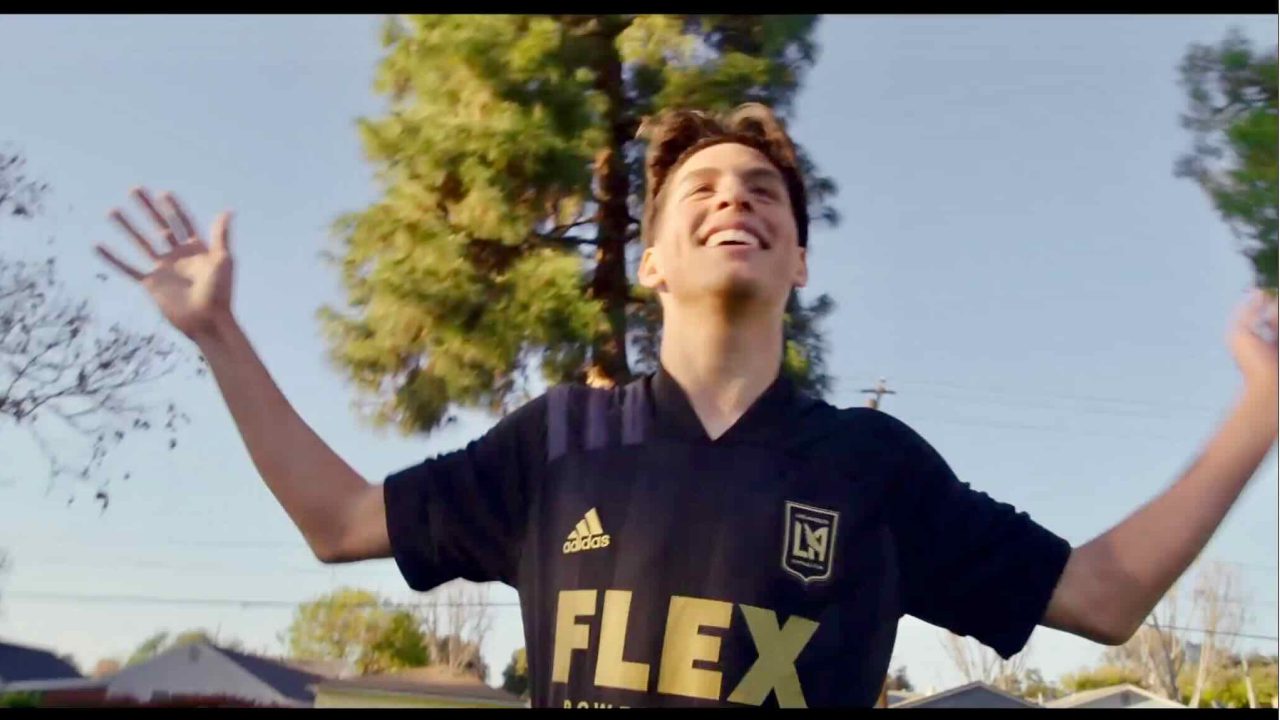 A person wearing a black soccer jersey with "FLEX" on the front extends their arms in joy outdoors with trees and blue sky in the background, capturing a perfect moment for content marketing.