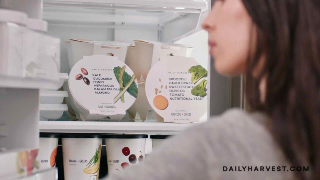 A health-conscious woman is looking into a refrigerator that contains neatly arranged Daily Harvest branded food containers. These plant-based meal delivery options highlight the convenience and nutrition of chef-crafted meals, including ready-to-eat smoothies, all promoting sustainability.