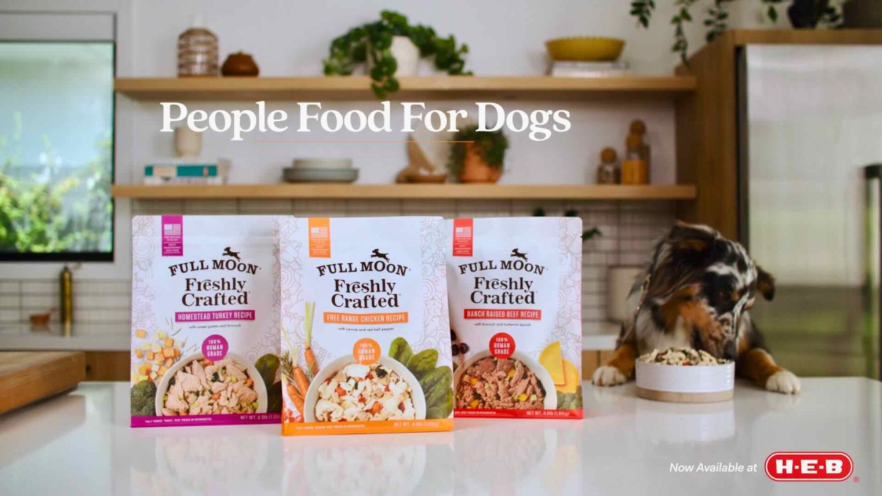 Three packages of Full Moon dog food are placed on a kitchen counter with a dog eating from a bowl in the background. Text above reads "People Food For Dogs" and the H-E-B logo is in the corner, making this scene look like an innovative television commercial.
