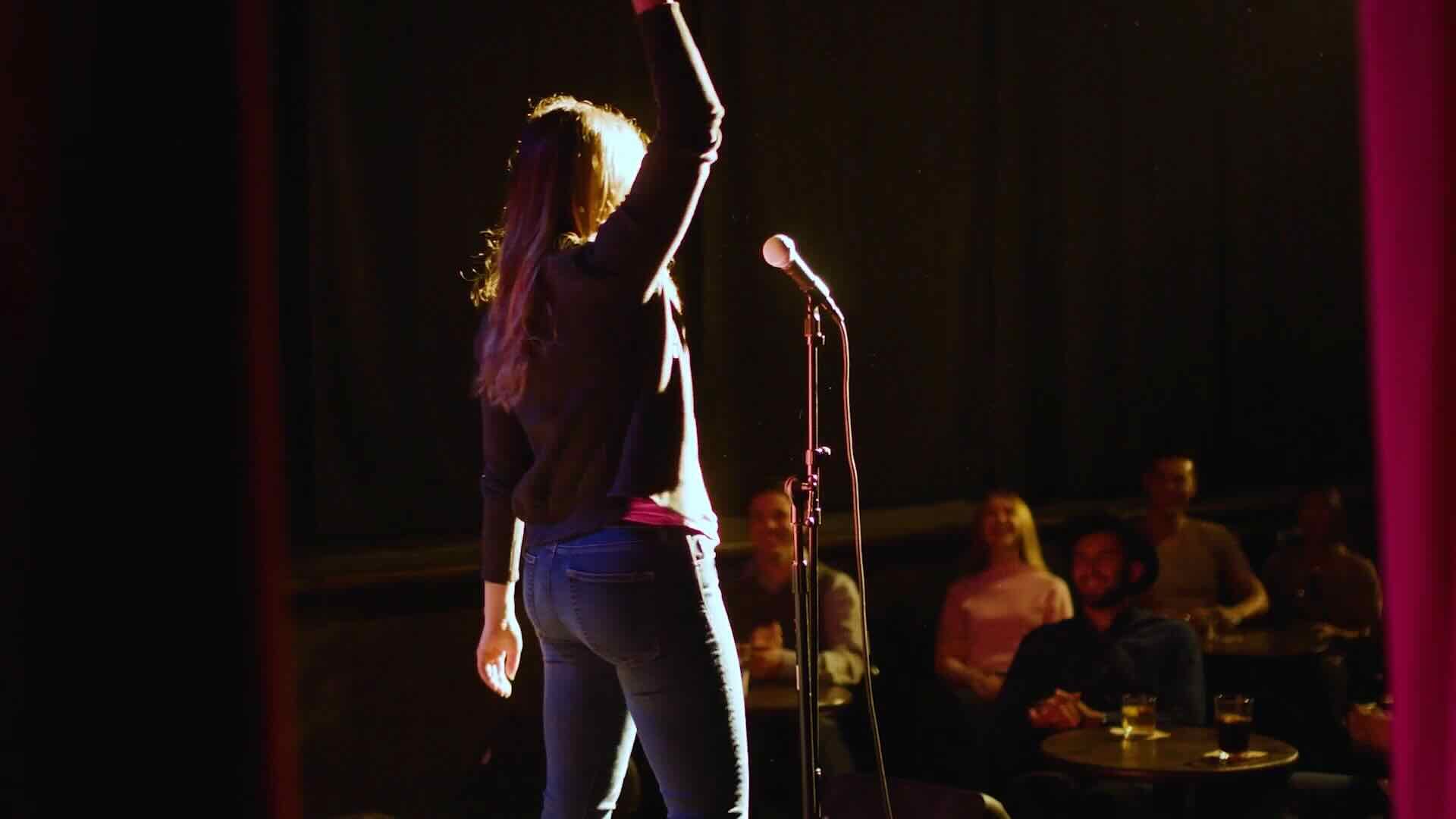 A person stands on stage with their arm raised, facing the audience, next to a microphone stand. Several spectators are seated and watching attentively, as if attending a captivating seminar on Medical Device marketing.