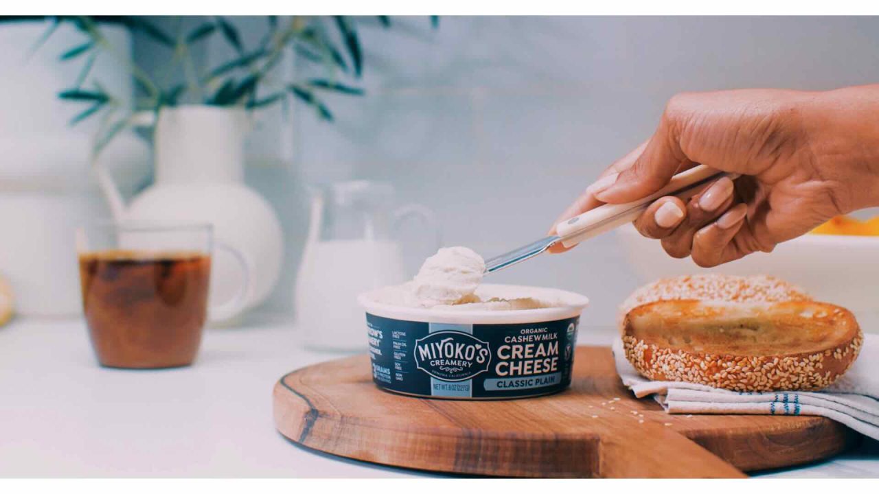 A hand holds a knife spreading Miyoko's cashew cream cheese onto a bagel on a wooden board, with iced coffee, a white vase, and a milk jug in the background – perfect for your next content marketing shot.