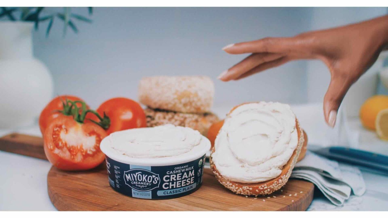 A hand spreads Miyoko's cashew cream cheese on a bagel, exemplifying effective content marketing. The background showcases tomatoes, a lemon, and bread rolls on a wooden board.