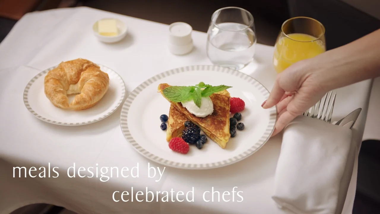 A hand placing a napkin beside a plate with French toast, berries, and mint garnish, next to a croissant, butter, water, and orange juice on a white tablecloth. Text reads: "Onboard dining options designed by celebrated chefs for Singapore Airlines' New York to Singapore direct flight.