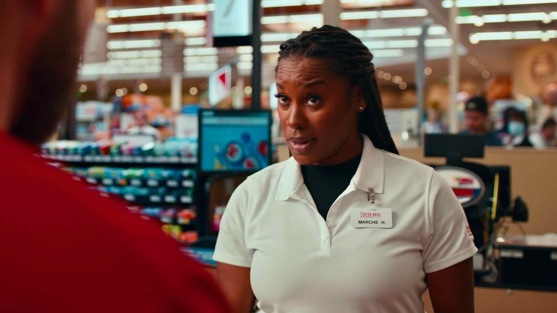 A woman in a white polo shirt with a name tag stands behind a counter in a Stater Bros. Markets store, attentively speaking to a customer about the supermarket's commitment to providing quality products and affordable shopping for busy families.