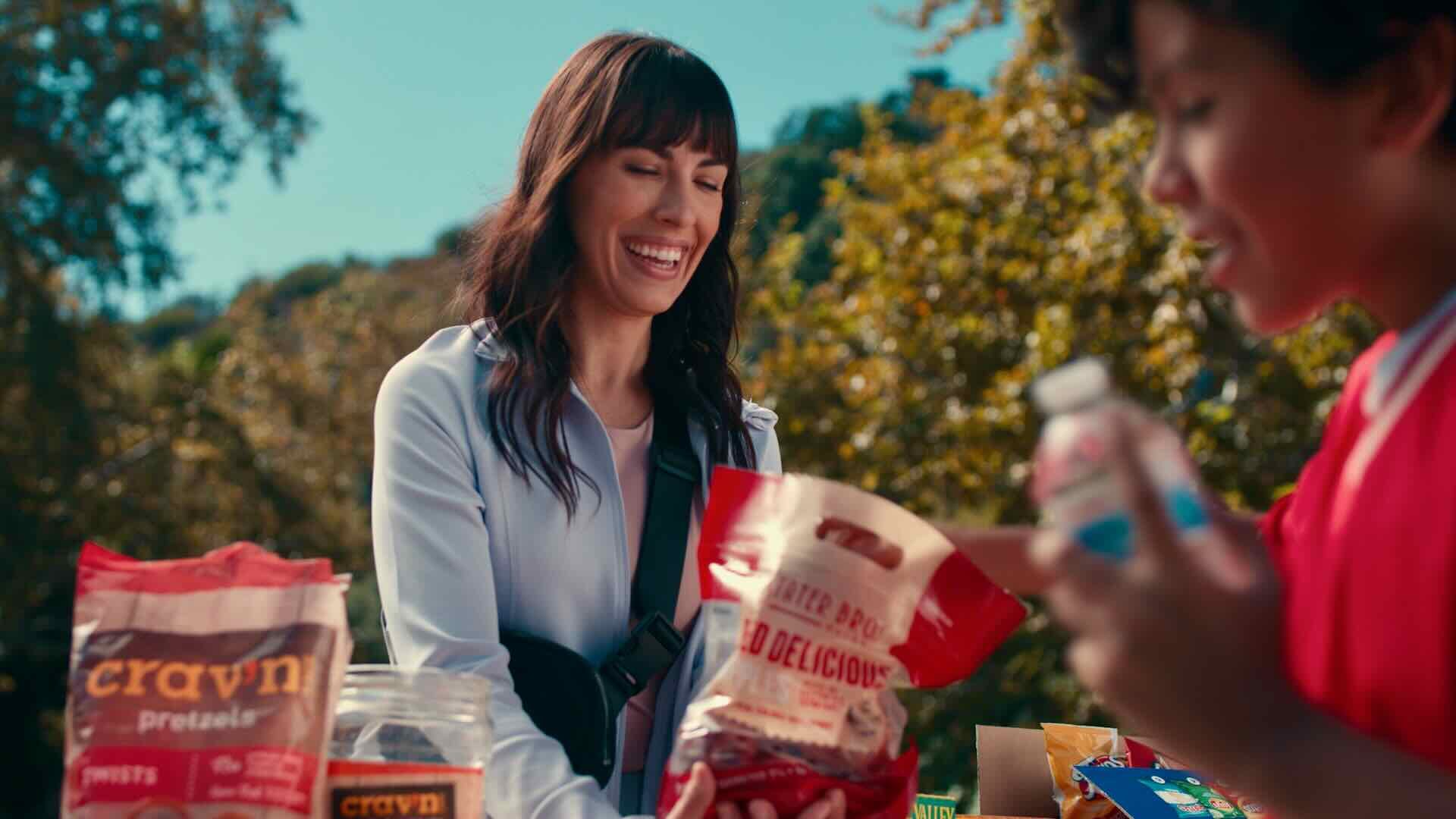 A woman smiles as she hands a bag of chips to a child at an outdoor snack stand filled with various foods and beverages, showcasing the variety and quality products that make Stater Bros. Markets a trusted provider for busy families in Southern California.